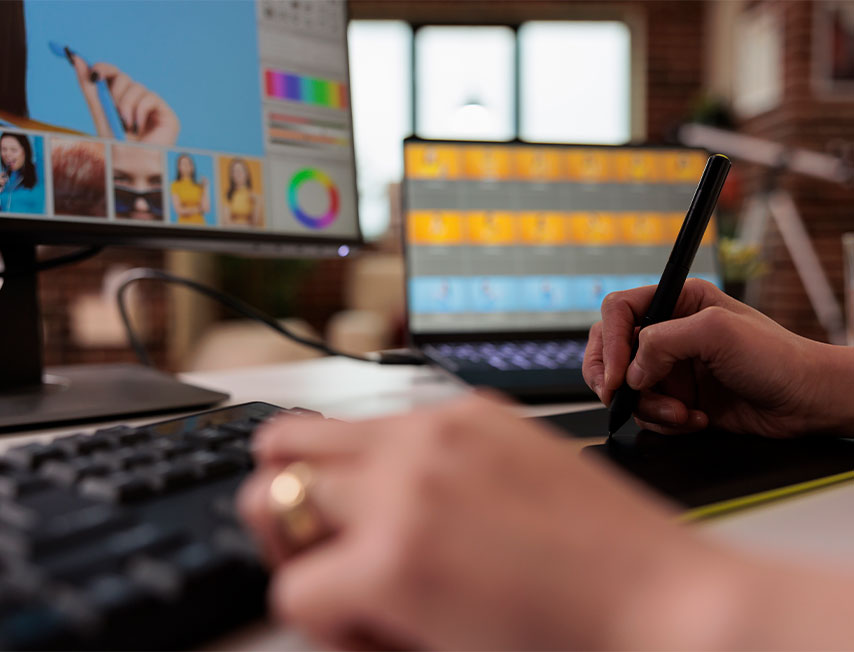 Imagem colorida, composta em um quarto ou studio, cujo a imagem principal é a mão de uma mulher, editando imagens em um aplicativo. Na composição da imagem há uma mesa digitalizadora, computador com o aplicativo de edição, e ao fundo desfocado um notebook com uma seleção de fotos.
