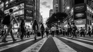 Pedestres cruzando uma faixa larga em avenida comercial movimentada de uma metrópole, em preto e branco, representando o fluxo de pessoas que forma a área de influência de uma loja