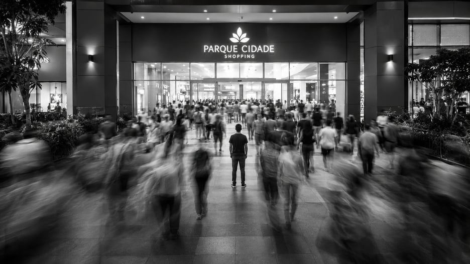 Multidão em movimento desfocado entrando em um shopping moderno à noite, com uma pessoa parada e nítida ao centro, em preto e branco, representando o fluxo real de visitantes de uma loja