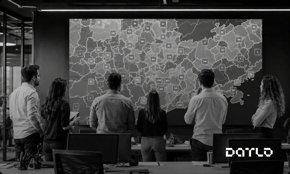 Equipe comercial de costas diante de um grande painel com o mapa de uma região do Brasil coberto por marcadores numerados de empresas, com áreas cheias e áreas vazias, representando a cobertura de mercado de uma operação B2B