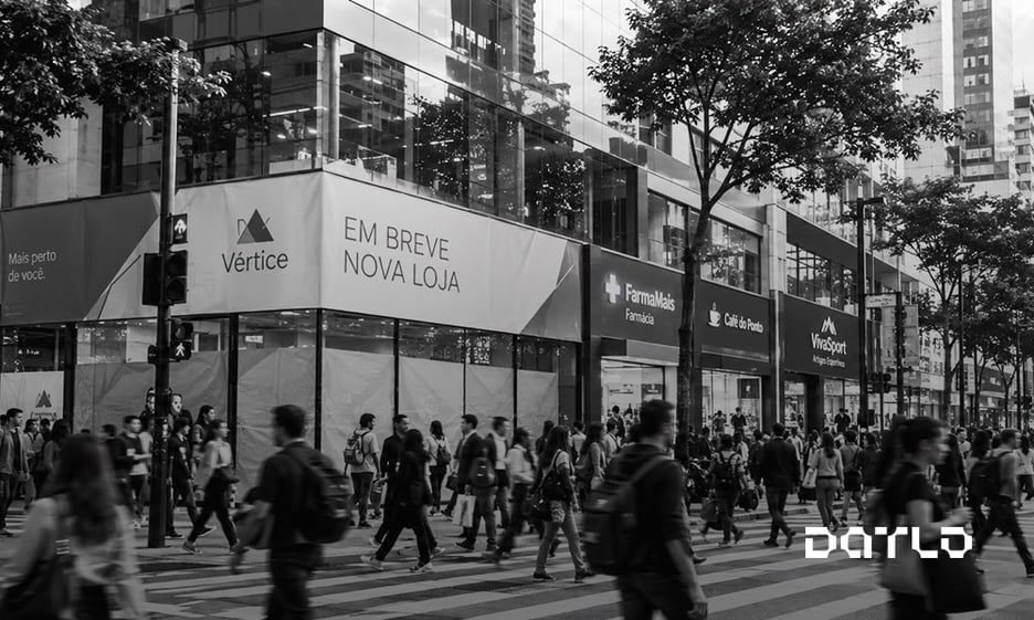 Pedestres cruzando uma avenida comercial movimentada diante de uma loja com o aviso de em breve nova loja, em preto e branco, representando a decisão de onde abrir uma nova unidade